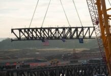 Intel Moves Forward at Ohio One With First Major Structural Truss Installation Arizona Fab 62 Tooling A large steel truss being lifted by a crane at Intel's Ohio One construction site, with an American flag attached.